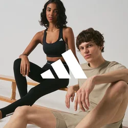 Due giovani adulti in abbigliamento sportivo sono seduti vicino a panche di legno in uno spazio interno minimalista, entrambi indossano scarpe da ginnastica Adidas.