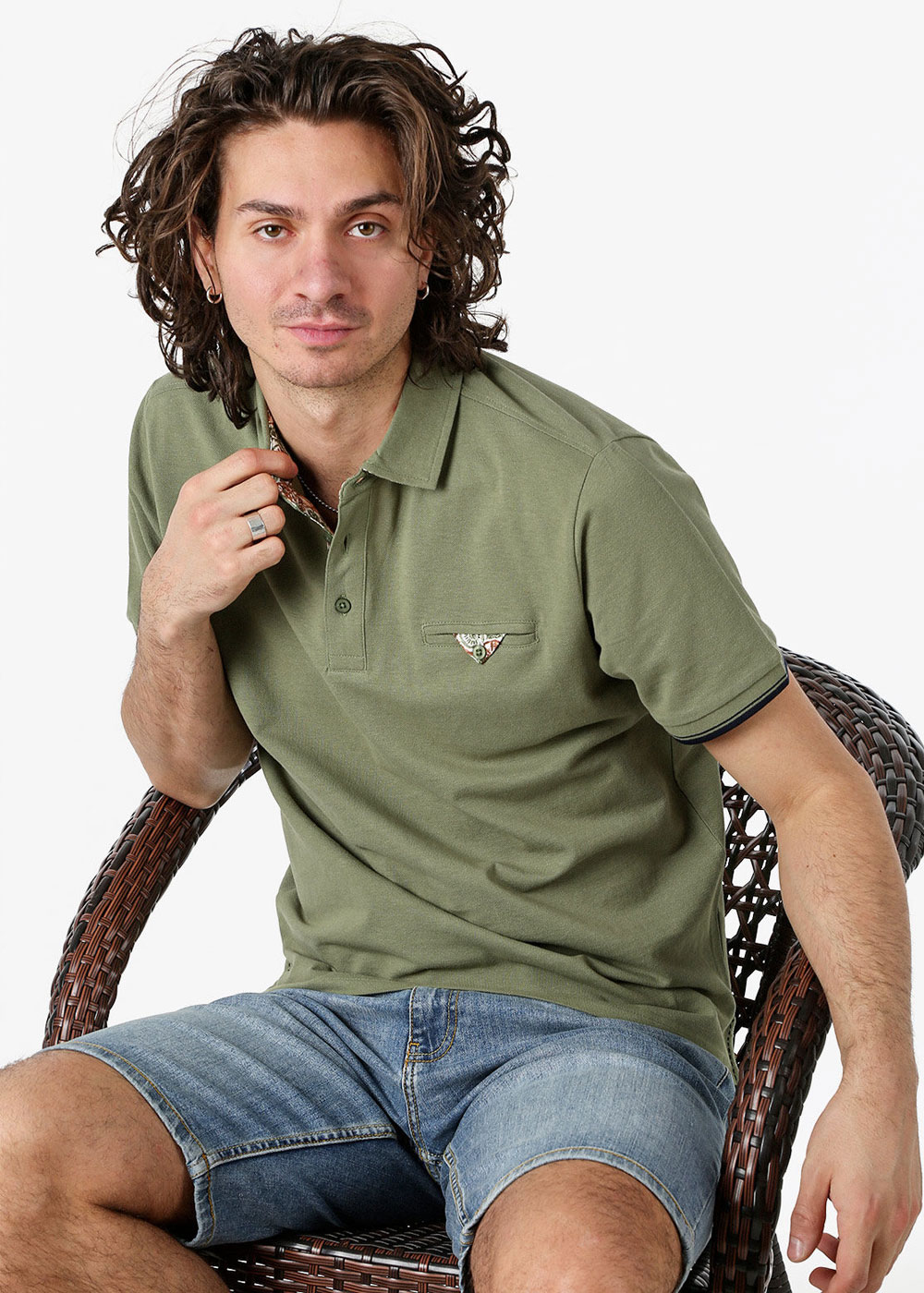 Young man model with curly hair sitting on a wicker chair, wearing an olive-green polo shirt and denim shorts in a casual summer outfit