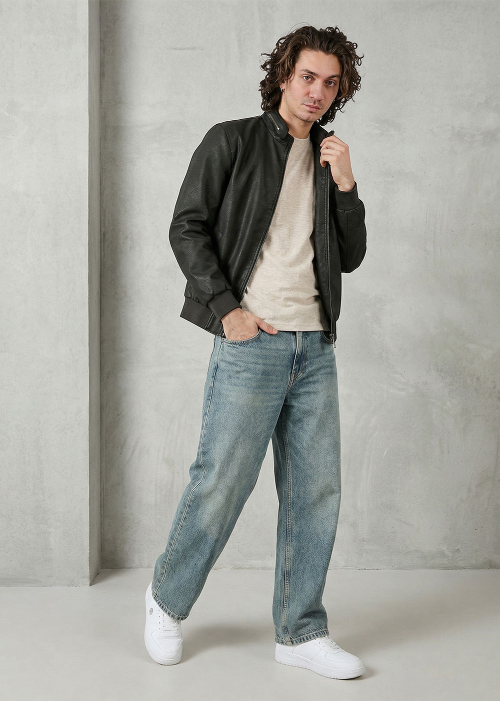 Man wearing a black leather bomber jacket, beige T‑shirt, loose blue jeans and white trainers standing against a concrete wall