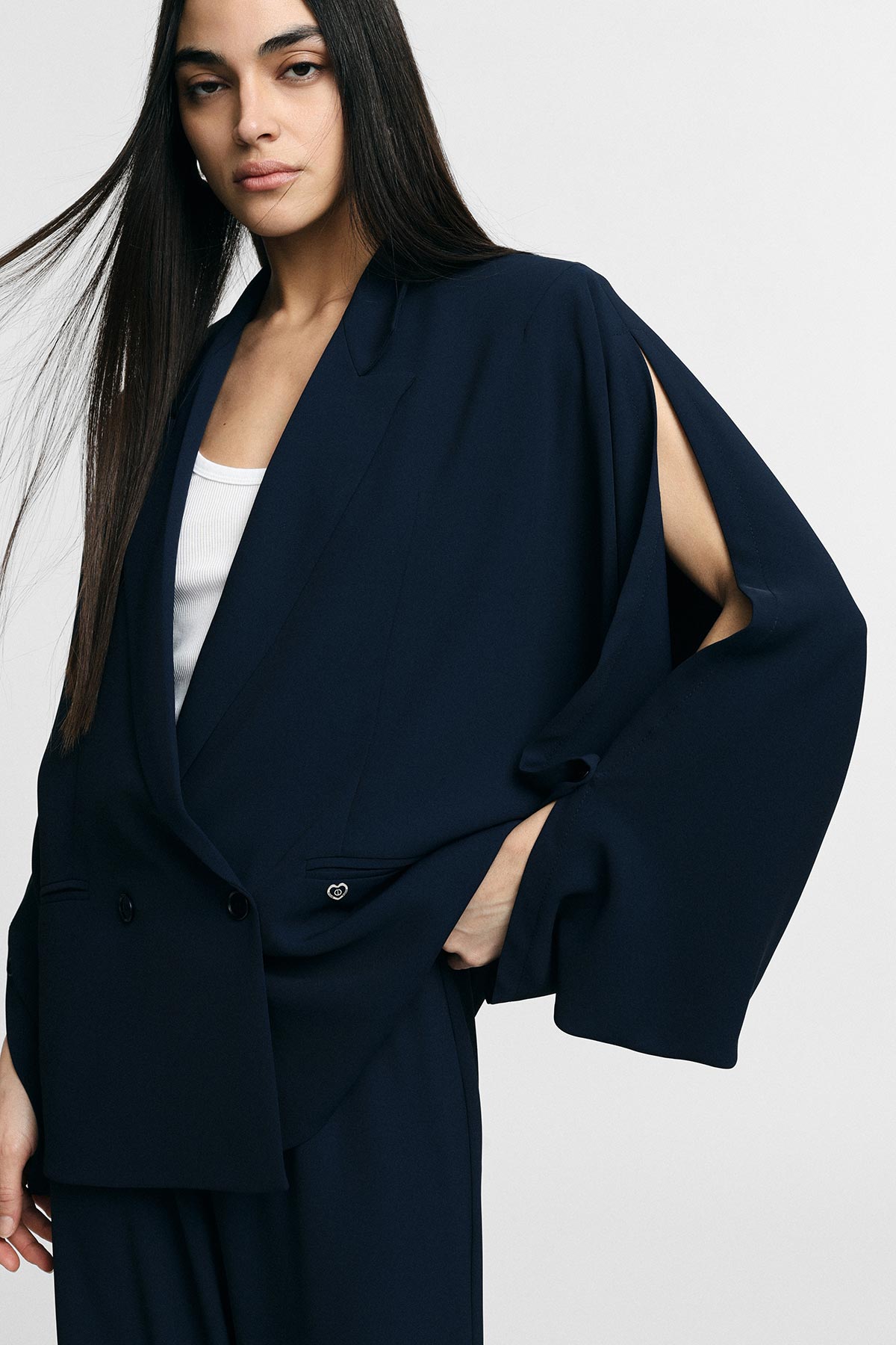 Woman wearing navy split-sleeve blazer and matching trousers with white ribbed top, hand in pocket, neutral studio background