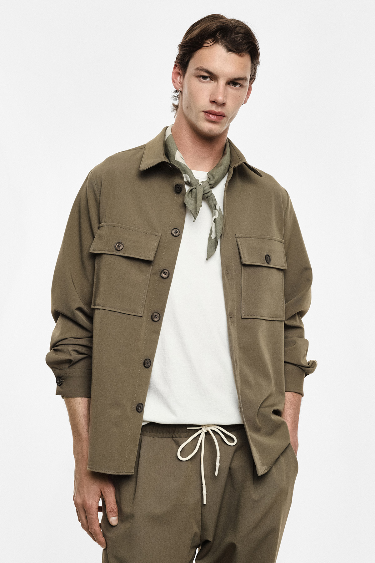 Young man modelling khaki overshirt and matching drawstring trousers with white T-shirt and neck scarf, hands in pockets