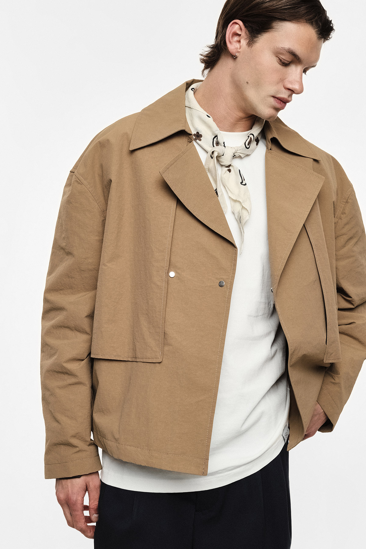 Male model wearing beige cropped jacket, cream patterned neck scarf and white top with navy trousers, looking down in a studio fashion shot
