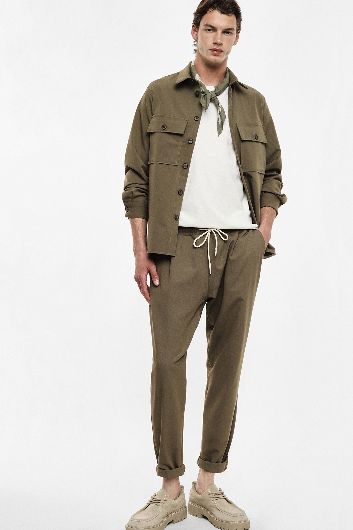 Full-length shot of a male model wearing khaki overshirt and drawstring trousers, white T-shirt, neck scarf and beige suede shoes on white background