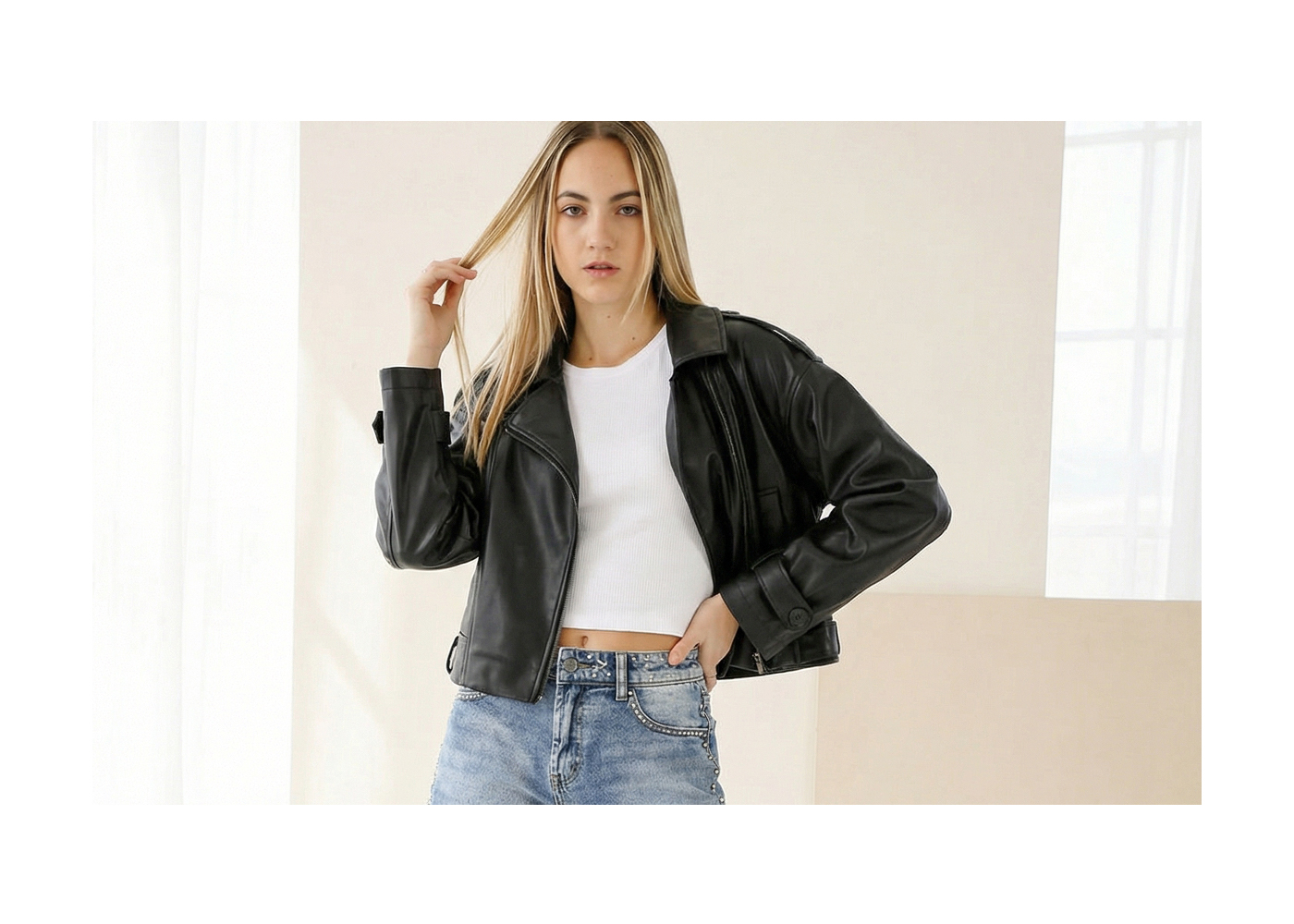 Woman in black leather jacket, white crop top and blue jeans posing indoors against a neutral background, hand in hair