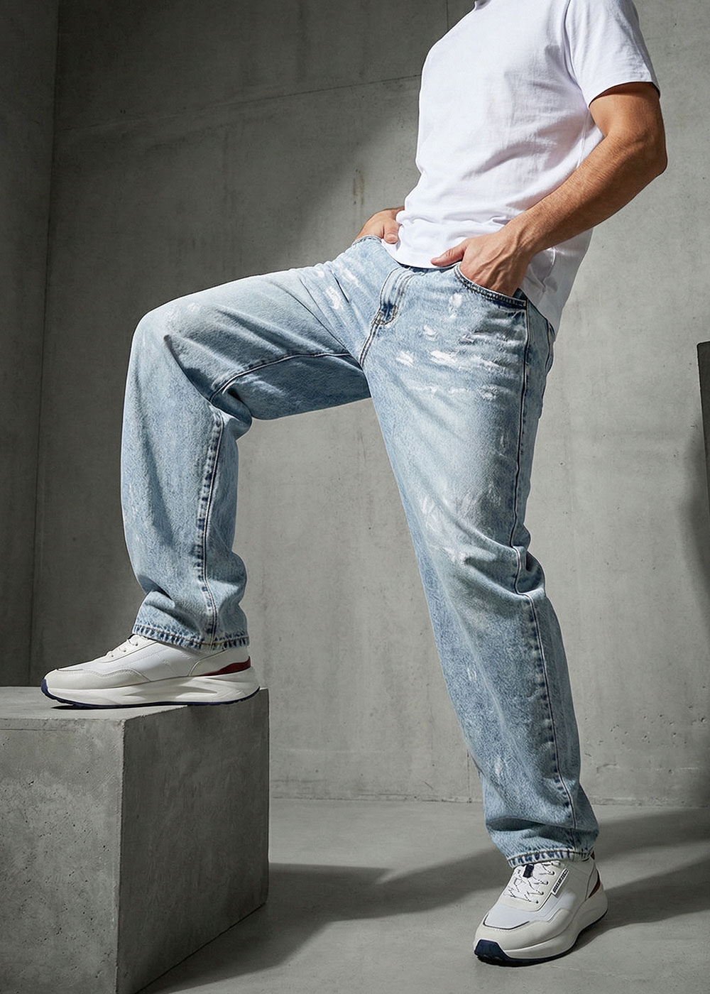 Man wearing white T-shirt, light-wash baggy jeans and white trainers, posing with one foot on a concrete block
