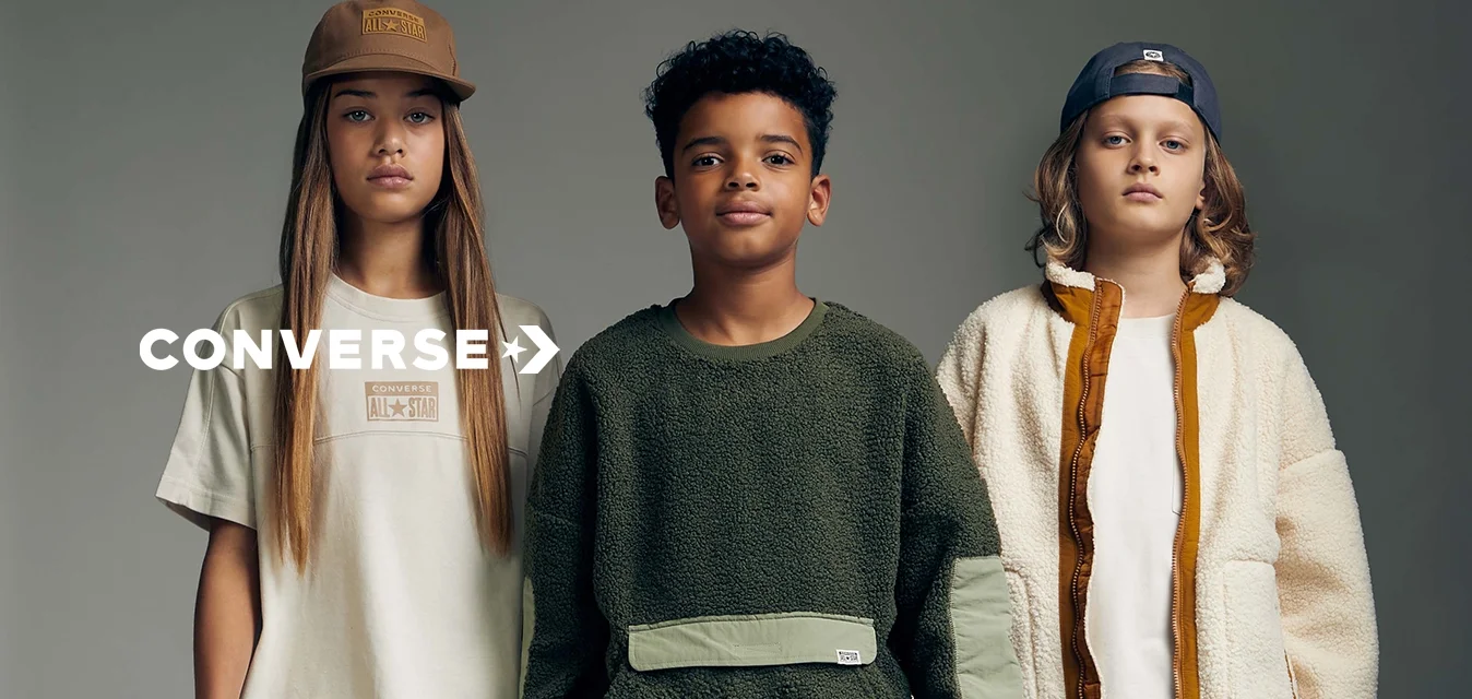 Tre bambini indossano outfit casual: una maglietta beige, una felpa verde oliva e una giacca crema, abbinate a pantaloni arancioni e verdi, tutti con sneakers.