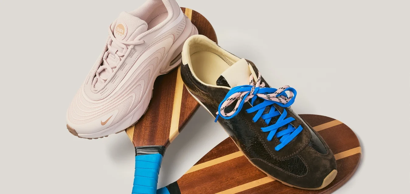 Due racchette da pickleball in legno con impugnature blu sovrapposte, sormontate da una sneaker bianca e una sneaker nera con lacci blu.