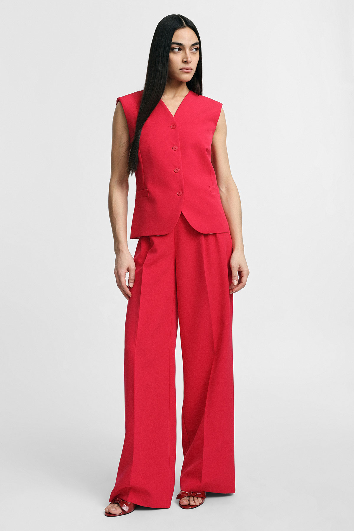 Woman wearing a red sleeveless tailored vest and wide-leg trousers, standing against a plain white background