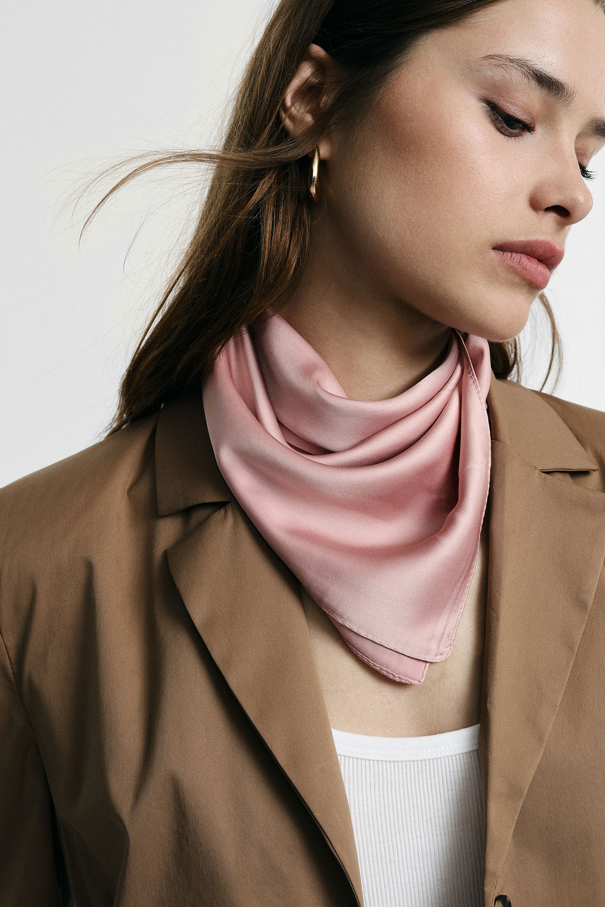 Close-up of a woman wearing a pale pink silk neck scarf, brown blazer, white top and gold hoop earring