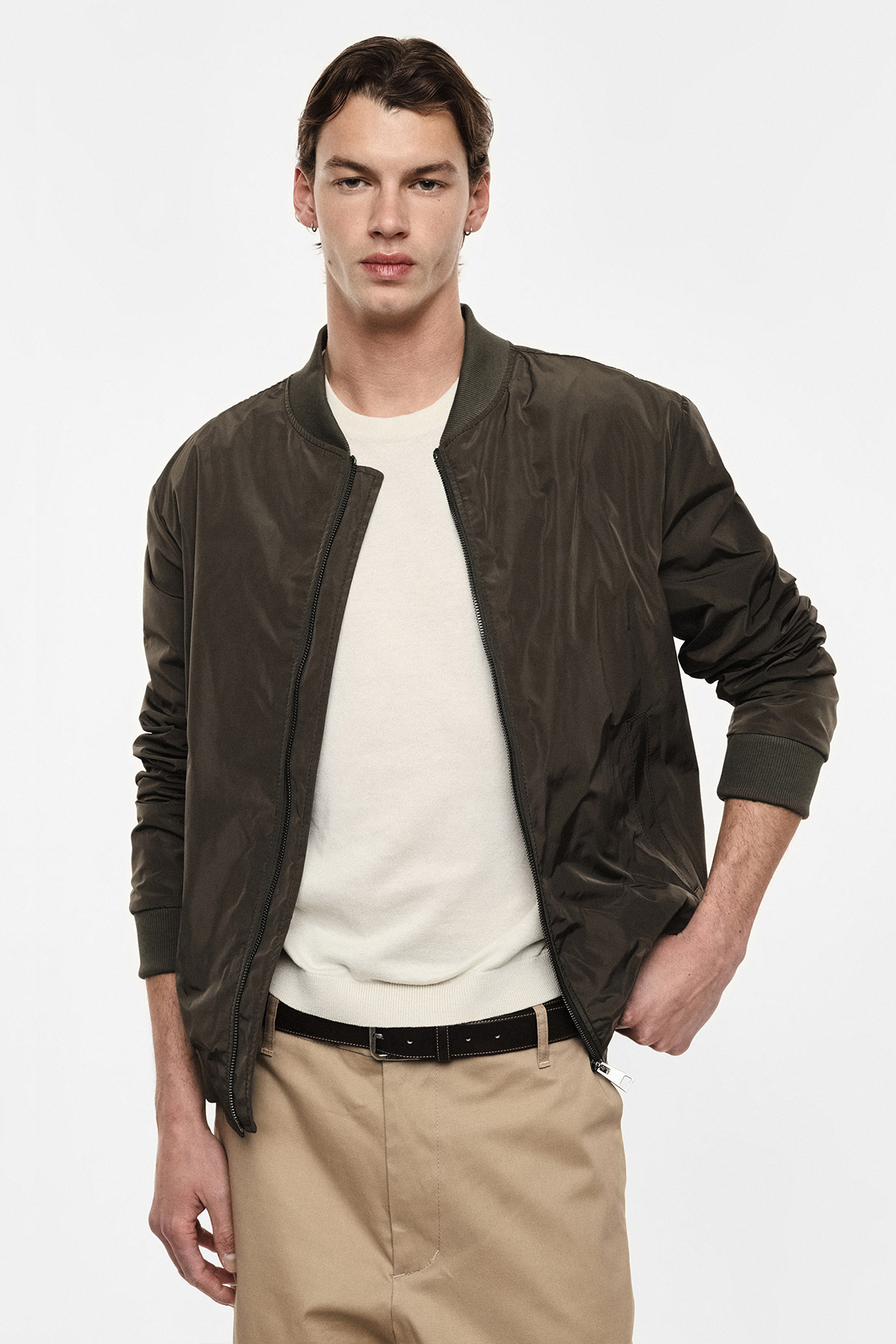 Young man wearing dark olive bomber jacket over cream sweater with beige chinos and black belt, hand in pocket on white background