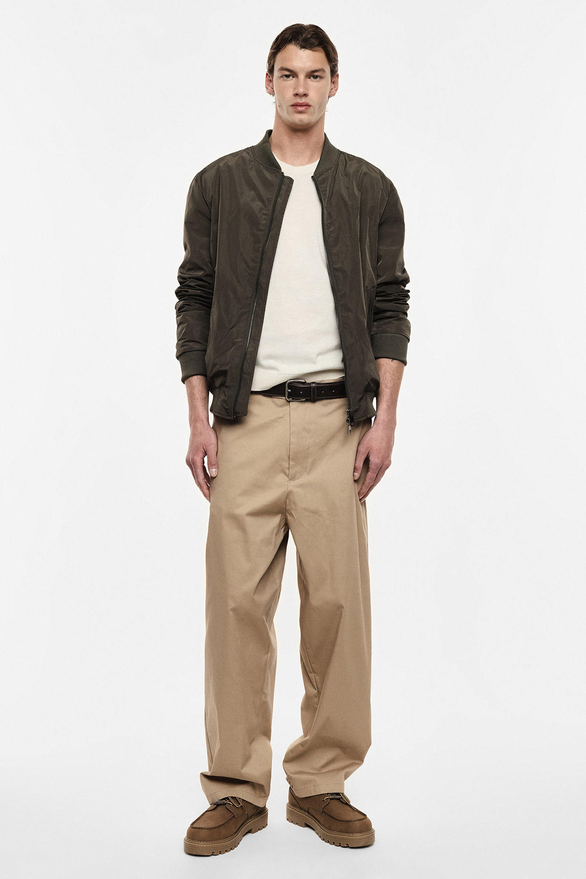 Male model wearing dark green bomber jacket, cream jumper, beige wide trousers, brown belt and shoes on white background