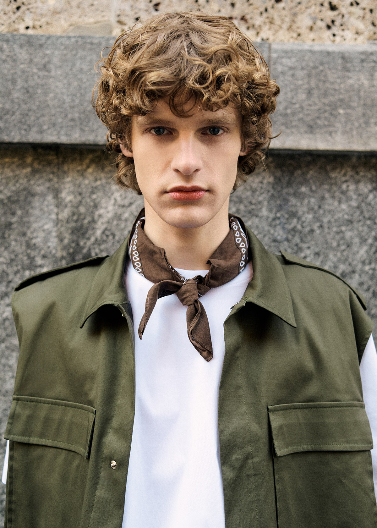 Young man with curly hair wearing green utility jacket, white T-shirt and brown neckerchief, standing by stone wall