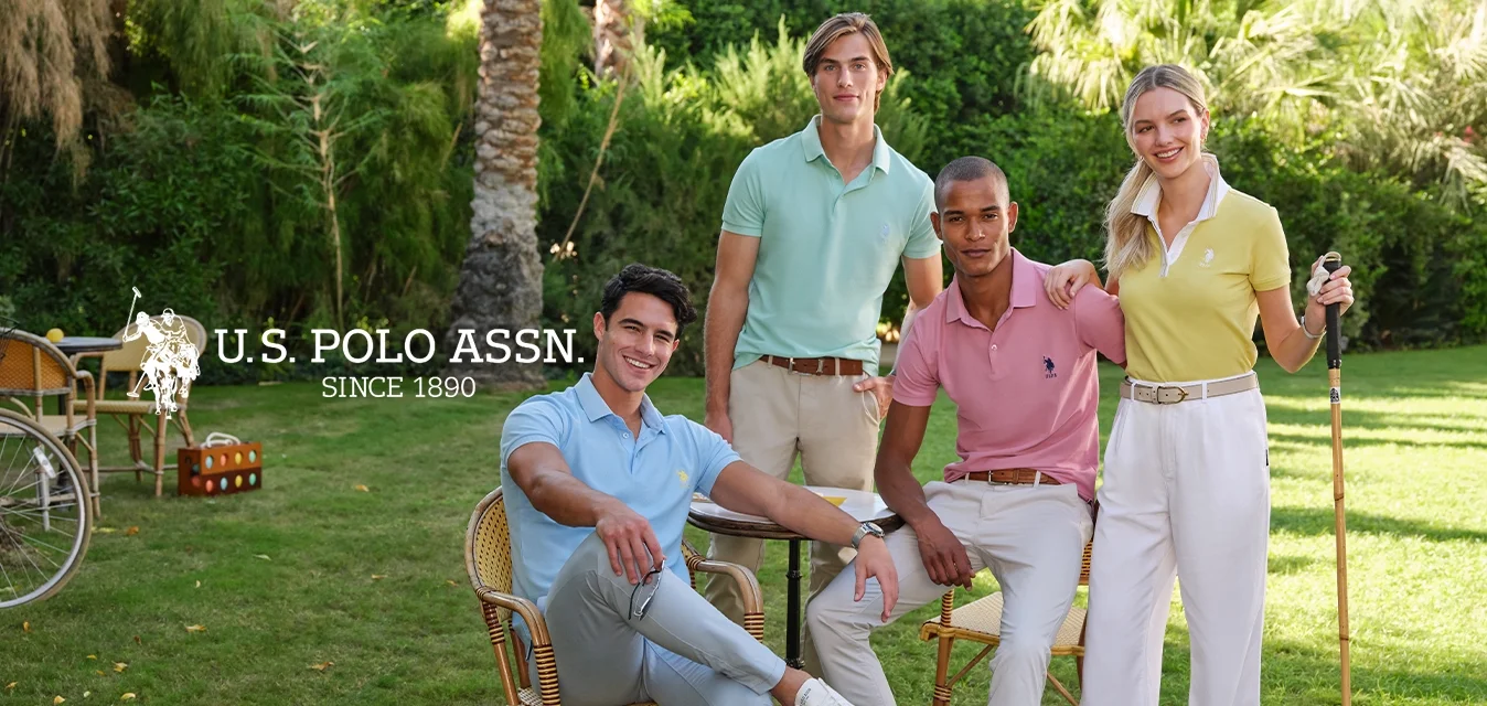 Quattro persone indossano polo pastello e pantaloni chiari, circondate da vegetazione. Una sedia in rattan e un martello di legno chiaro sono nelle vicinanze.