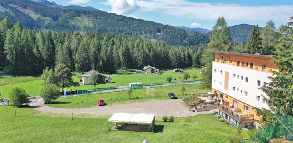 Alpine lodge beside a green meadow and pine forest with mountains in the background, small parking area and a tented gazebo