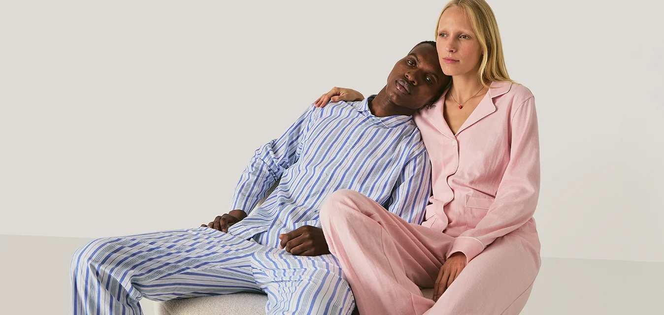 Un uomo in pigiama a righe blu poggia la testa sulla spalla di una donna che indossa un pigiama rosa, seduti vicini su un blocco beige.