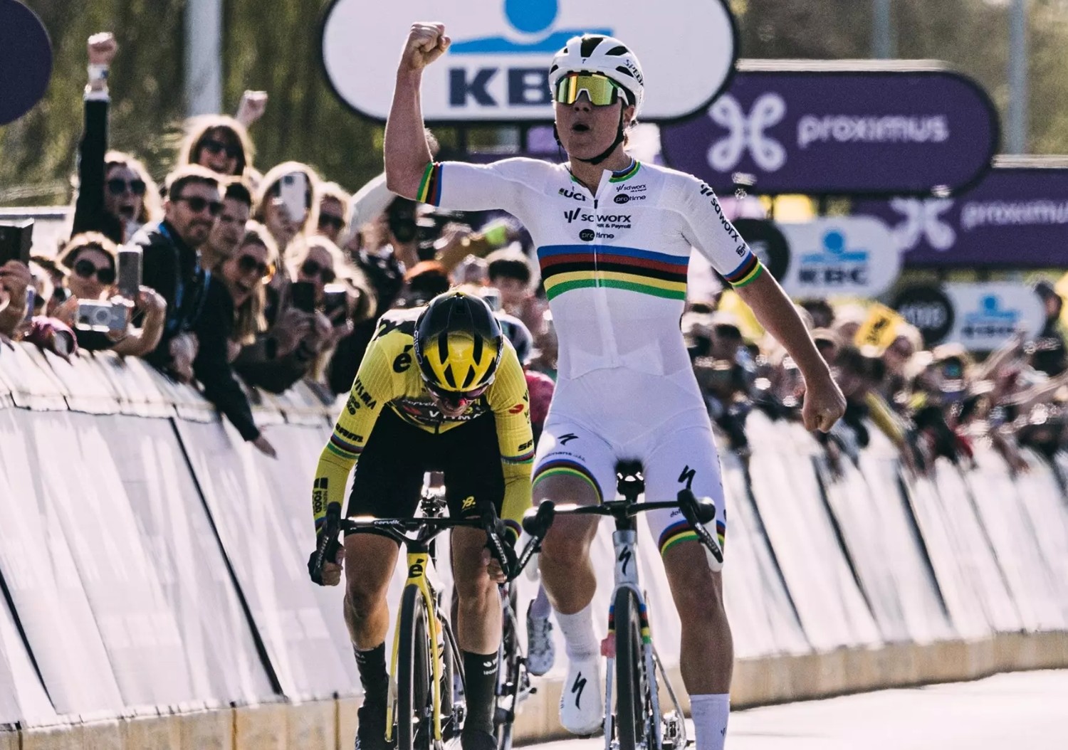 Cyclist in white rainbow jersey raising a fist to celebrate a sprint win, rival cyclist and cheering crowd behind