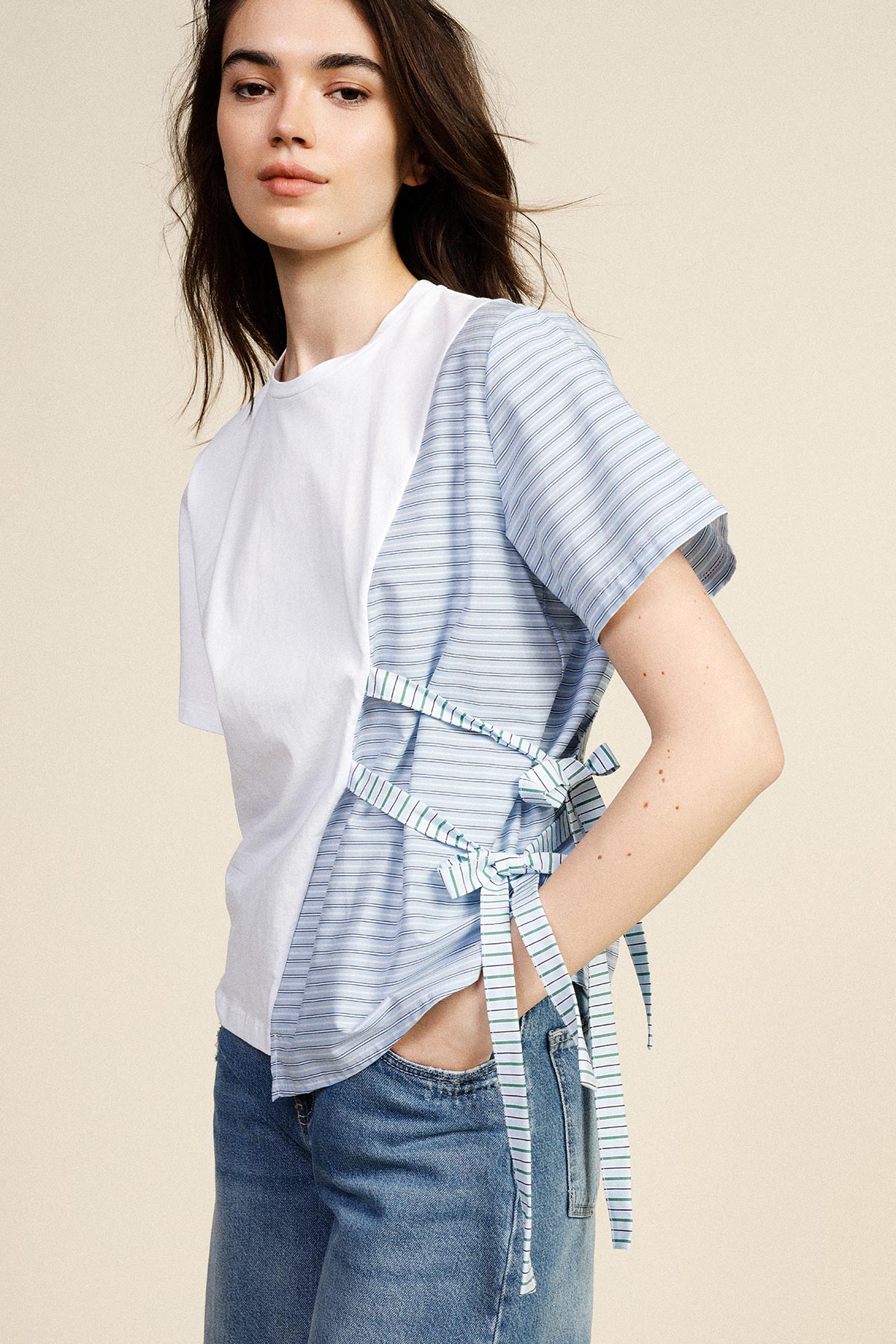 Young woman wearing white and blue striped short-sleeve top with side tie details and blue jeans against neutral background