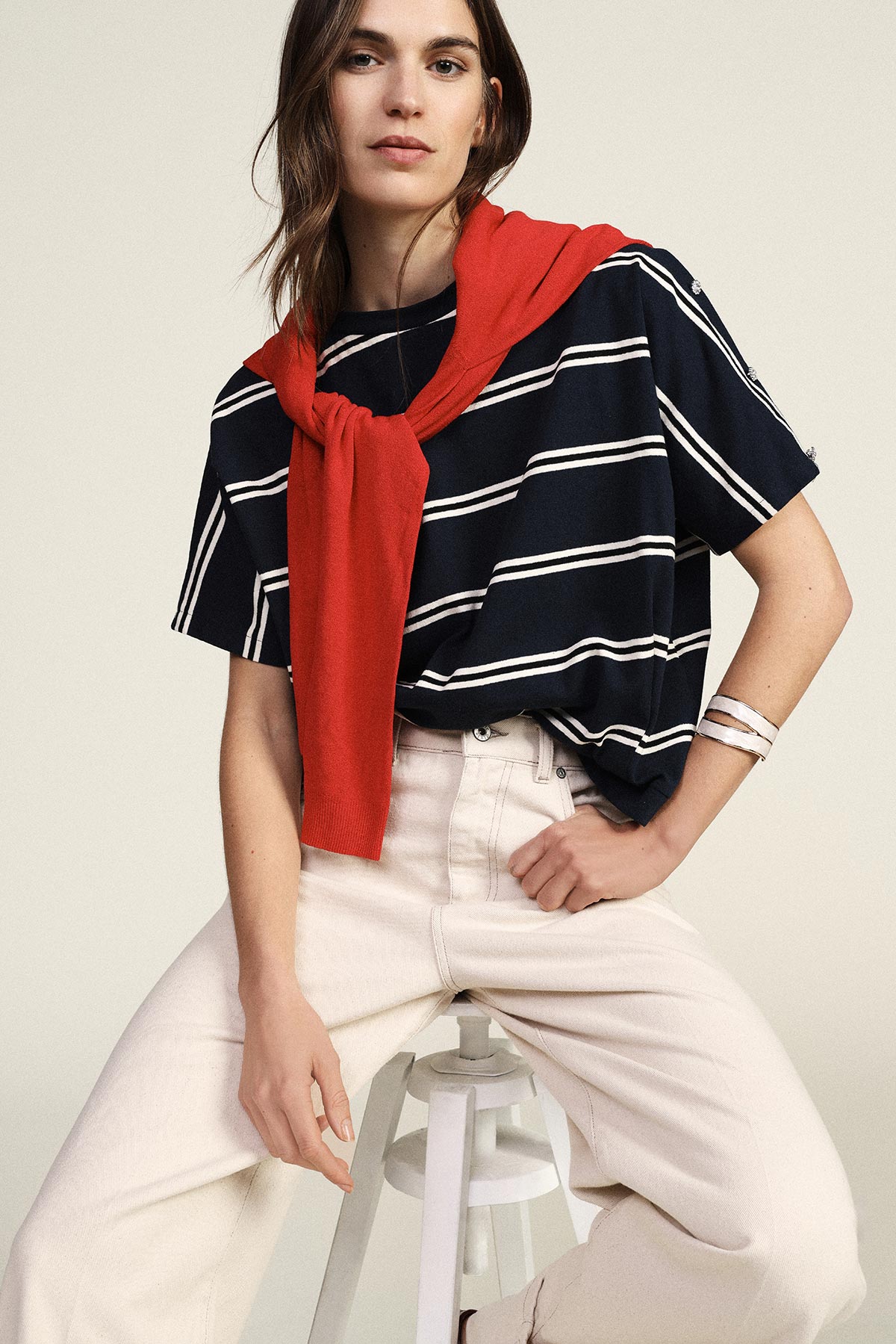 Woman seated on stool wearing navy striped T‑shirt, red sweater draped over shoulders and cream jeans