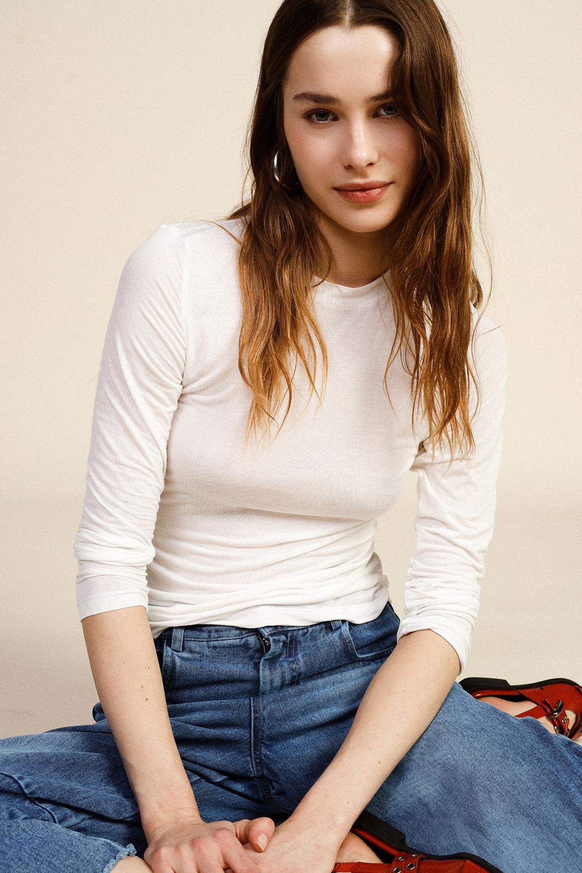 Young woman in a white long-sleeve top and blue jeans sitting cross-legged against a neutral background