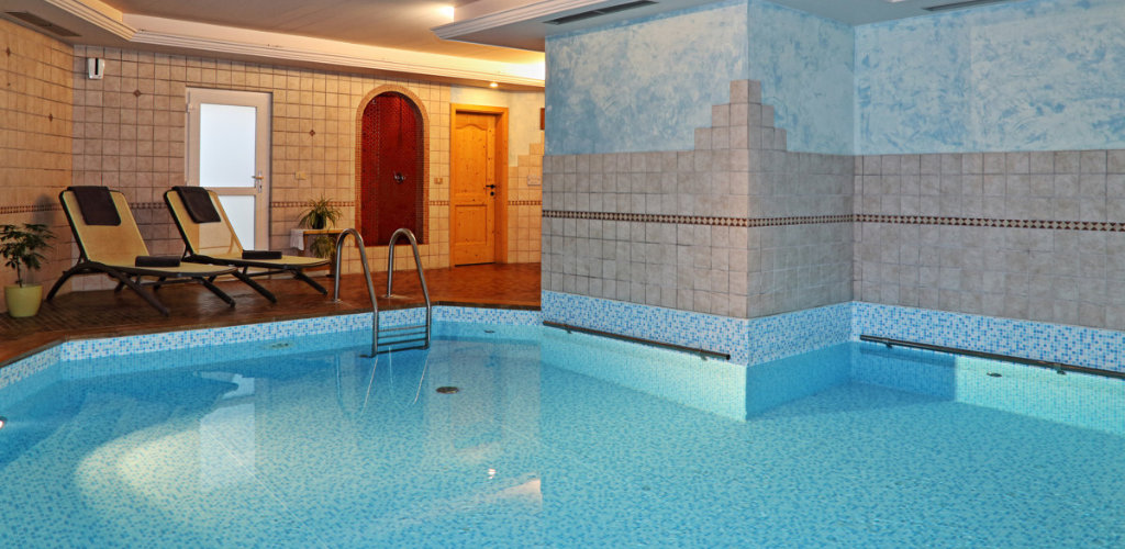 Indoor tiled swimming pool and spa area with metal ladder, two reclining loungers and wooden doors in a cosy wellness room
