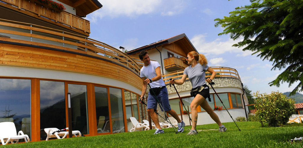Couple Nordic walking with poles on lawn outside a wooden alpine chalet hotel at a mountain resort