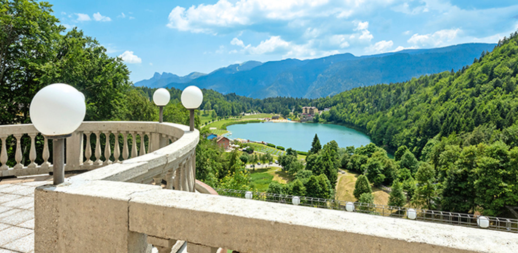 Stone terrace with round globe lamps overlooking a turquoise mountain lake, lush green forest and distant blue peaks under a sunny sky