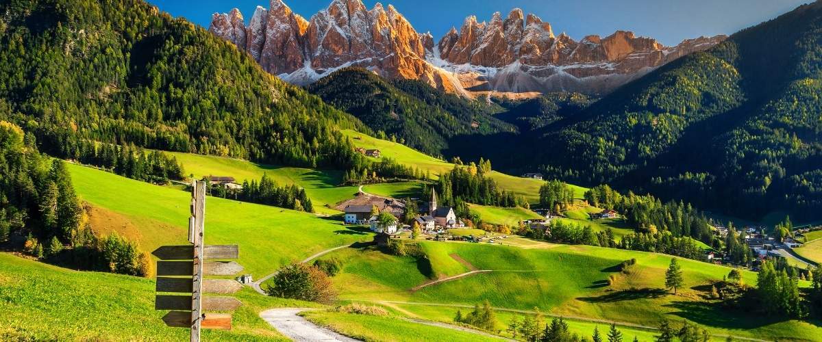 Wooden signpost by a winding path leading to an alpine village amid green pastures and rugged sunlit mountains