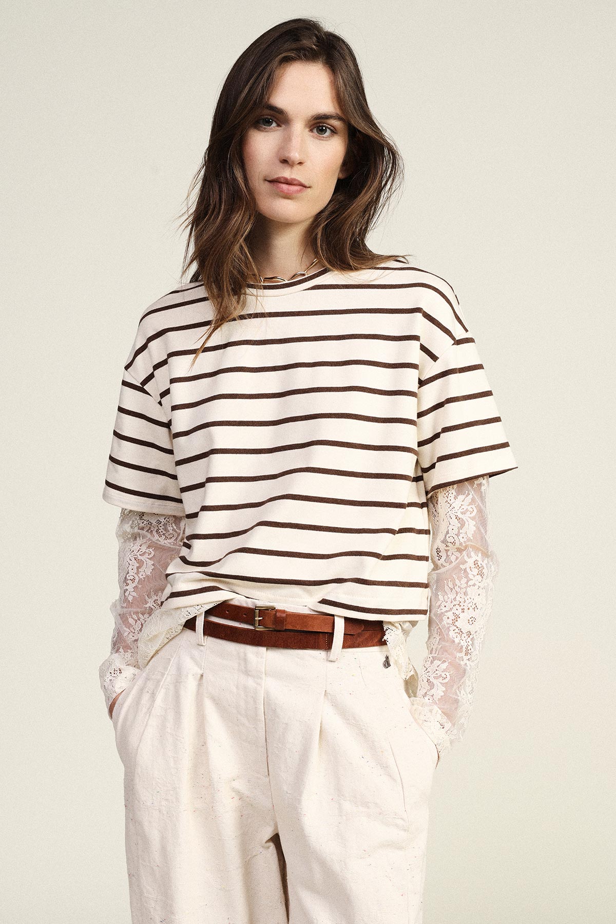 Young woman wearing cream and brown striped T‑shirt layered over white lace sleeves, cream trousers and brown belt, hands in pockets