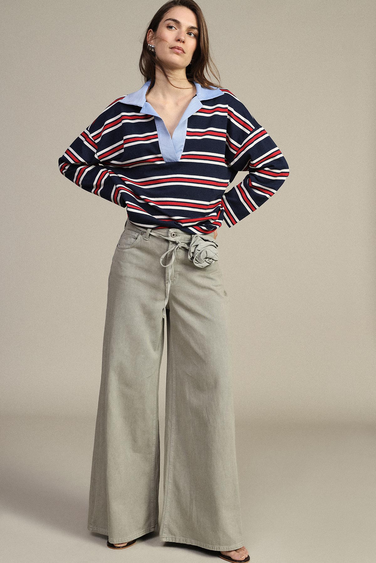 Model wearing navy, red and white striped rugby shirt with light blue collar and high-waisted wide-leg beige trousers, hands on hips