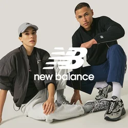 Donna con cappello grigio, giacca nera e pantaloni della tuta grigi siede accanto a un uomo con giacca nera e pantaloni blu, entrambi indossano sneakers grigie su sfondo semplice.