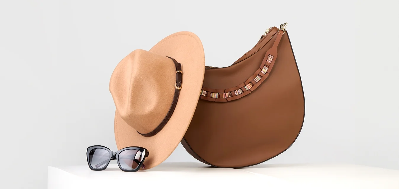 Borsa in pelle marrone con forma curva e dettagli decorativi, abbinata a un cappello fedora beige e occhiali da sole neri a forma di occhio di gatto su un piedistallo bianco.