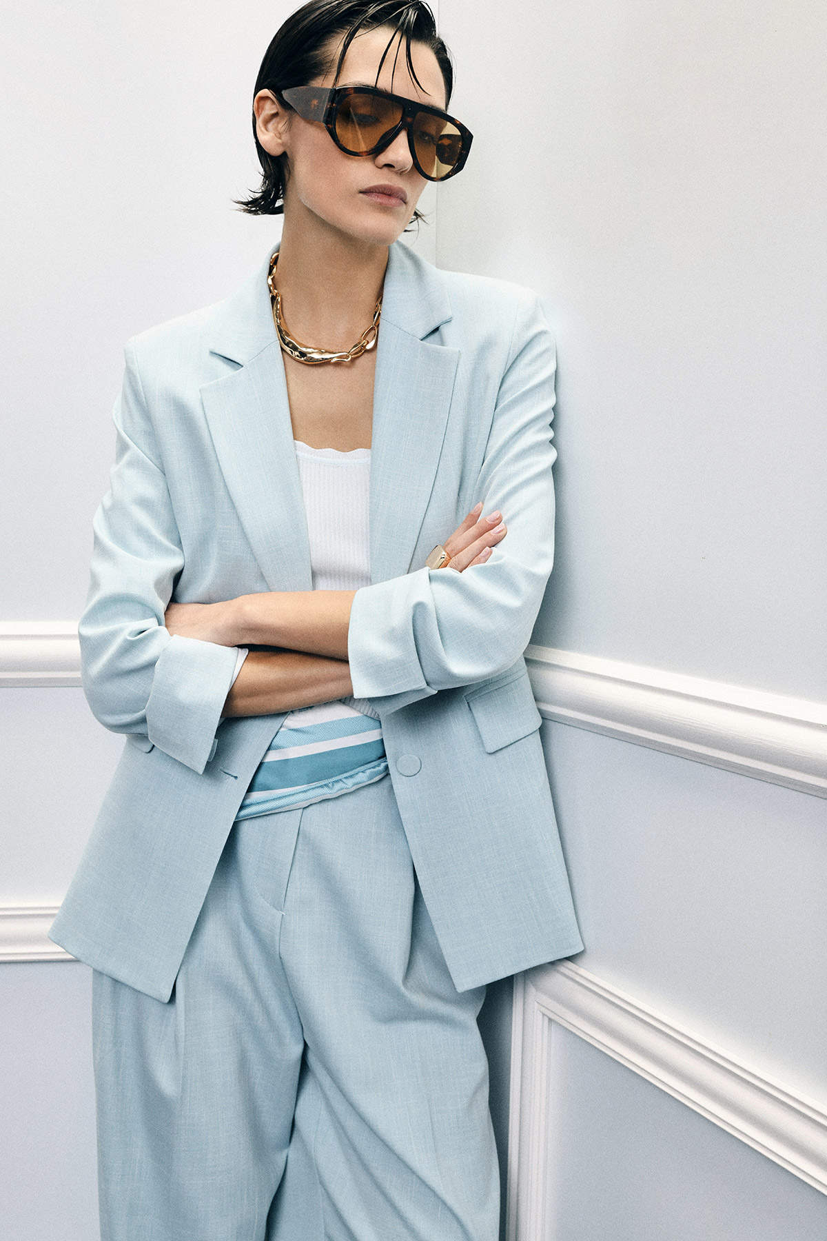 Woman wearing pastel blue tailored suit with oversized blazer, top, chunky gold chain and tinted sunglasses, arms crossed