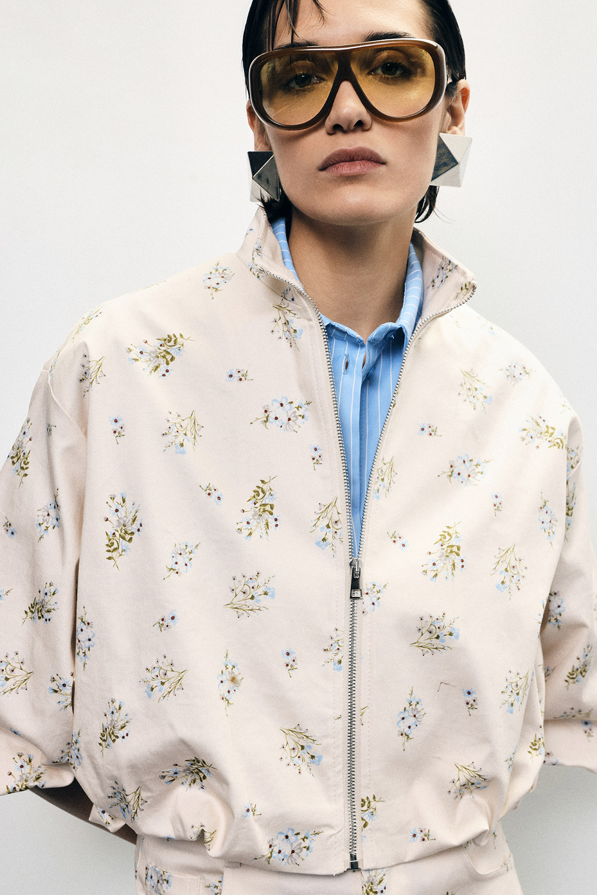 Fashion model wearing cream floral zip-up jacket, blue shirt, oversized amber sunglasses and silver geometric earrings
