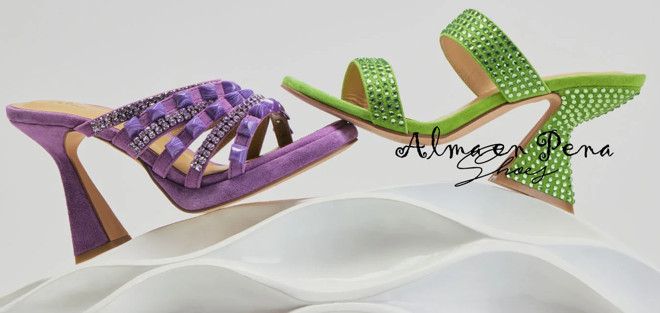 Sandali con tacco viola e verde con strass, esposti in equilibrio su una superficie astratta bianca e ondulata contro uno sfondo semplice.