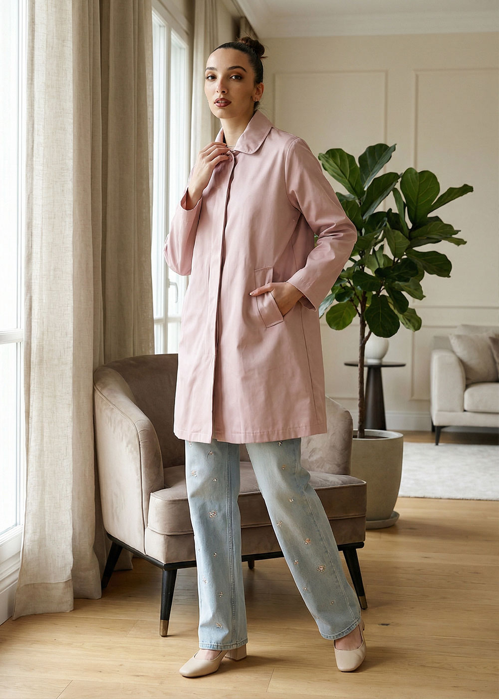 Woman wearing a pink coat and embroidered jeans standing by a window in a stylish living room, posing beside an armchair and plant