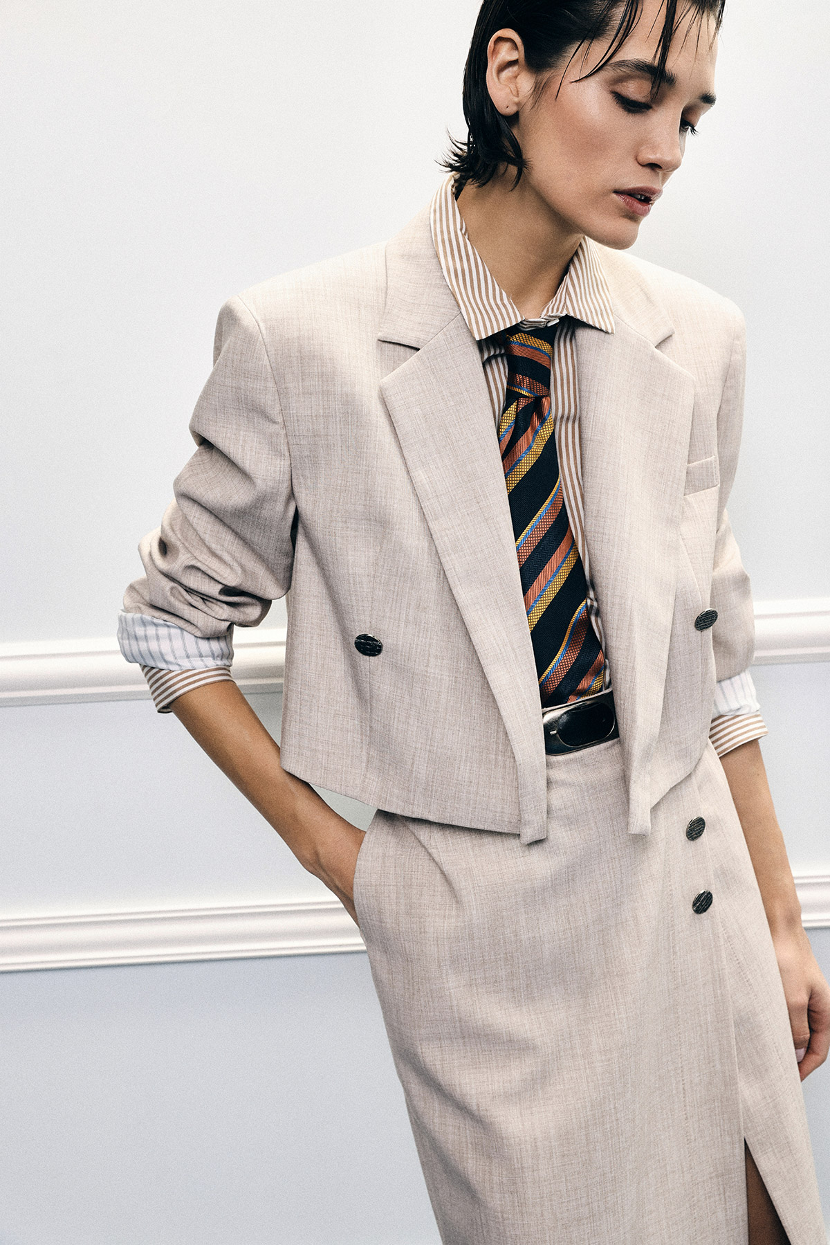 Model in a light beige cropped blazer and skirt with striped shirt and colourful tie, hand in pocket, looking down