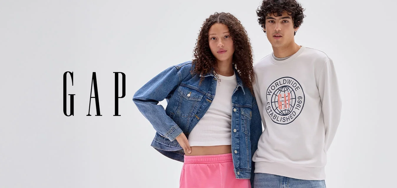 Due giovani adulti in piedi vicino, la donna indossa pantaloni della tuta rosa e giacca di jeans, l'uomo una felpa chiara e jeans, sfondo grigio neutro.