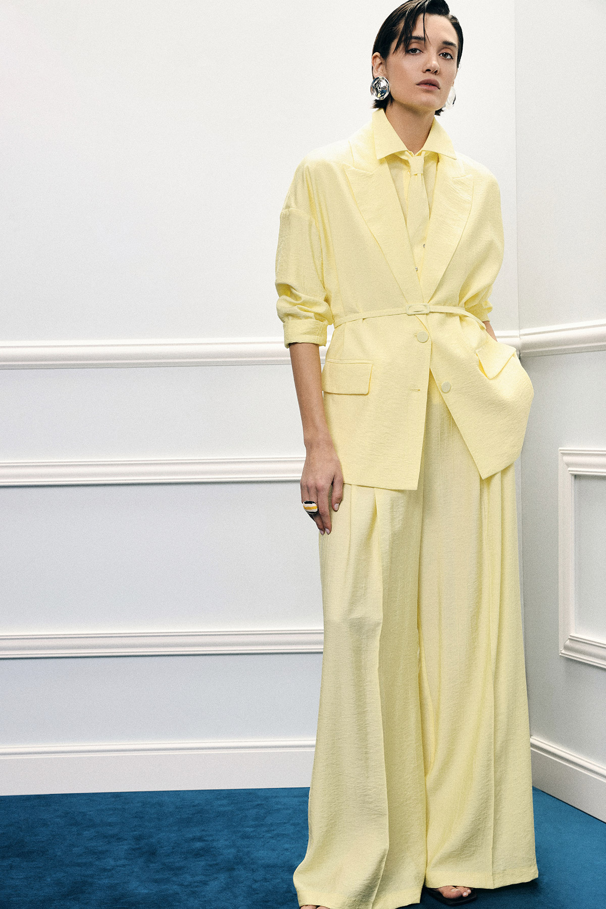 Woman in pale yellow oversized blazer and wide‑leg trousers, statement earrings, standing by white wall and blue carpet