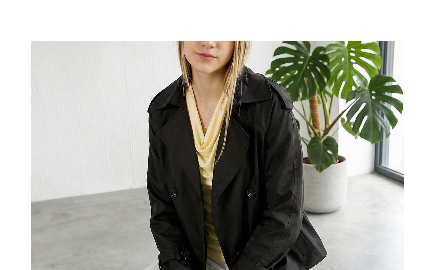 Woman wearing black trench coat over yellow cowl-neck top seated in bright modern room beside potted monstera plant