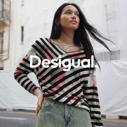 Donna con capelli lunghi e scuri che indossa un maglione a righe multicolore e una gonna di jeans, in piedi in una strada della città con edifici storici.