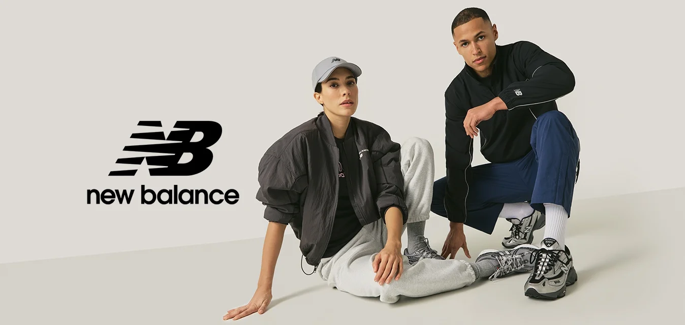 Donna con cappello grigio, giacca nera e pantaloni della tuta grigi siede accanto a un uomo con giacca nera e pantaloni blu, entrambi indossano sneakers grigie su sfondo semplice.