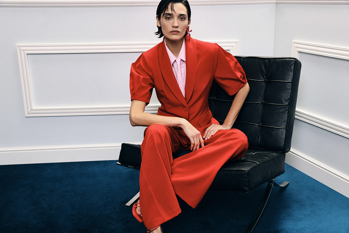 Woman wearing a red tailored suit and pale pink blouse seated on a black leather chair in a minimalist room