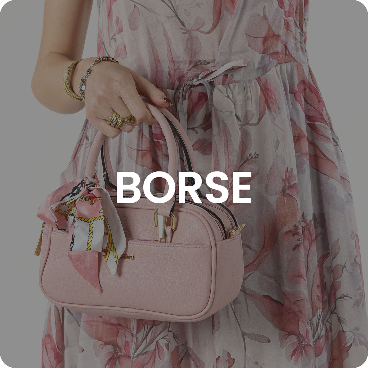 Woman holding small pale pink leather handbag with silk scarf tied to handle, wearing floral summer dress and gold jewellery