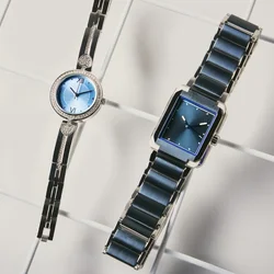 Due orologi da polso eleganti con quadranti blu e cinturini in metallo, uno rotondo con dettagli a forma di cuore, l'altro rettangolare, esposti su una superficie a griglia bianca.