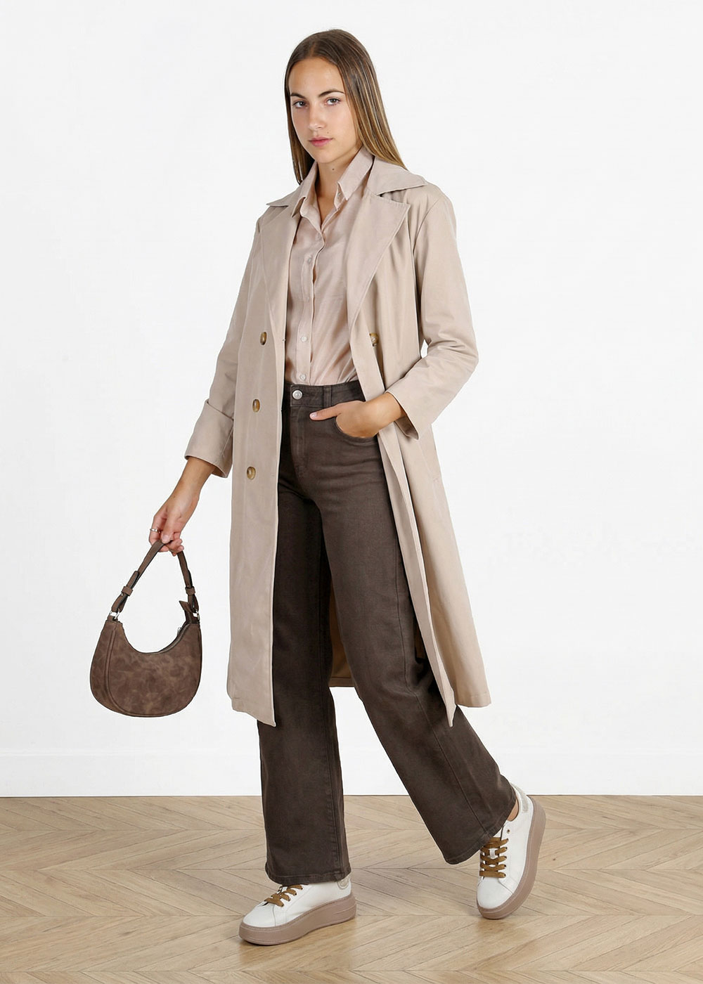 Woman in beige trench coat, pale blouse and dark brown wide-leg jeans, holding small brown suede bag and white trainers