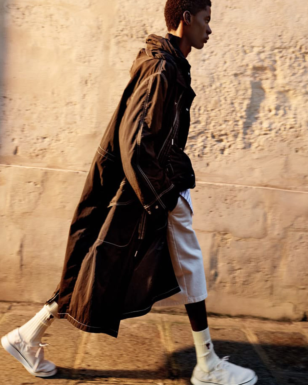 Profile of a person walking past a sunlit stone wall wearing a long black trench coat, beige knee-length shorts and white trainers