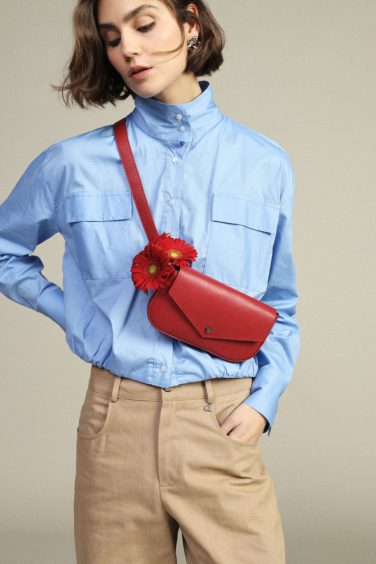Woman in light-blue high-collar shirt and beige trousers wearing a red leather crossbody bag with red gerbera flowers