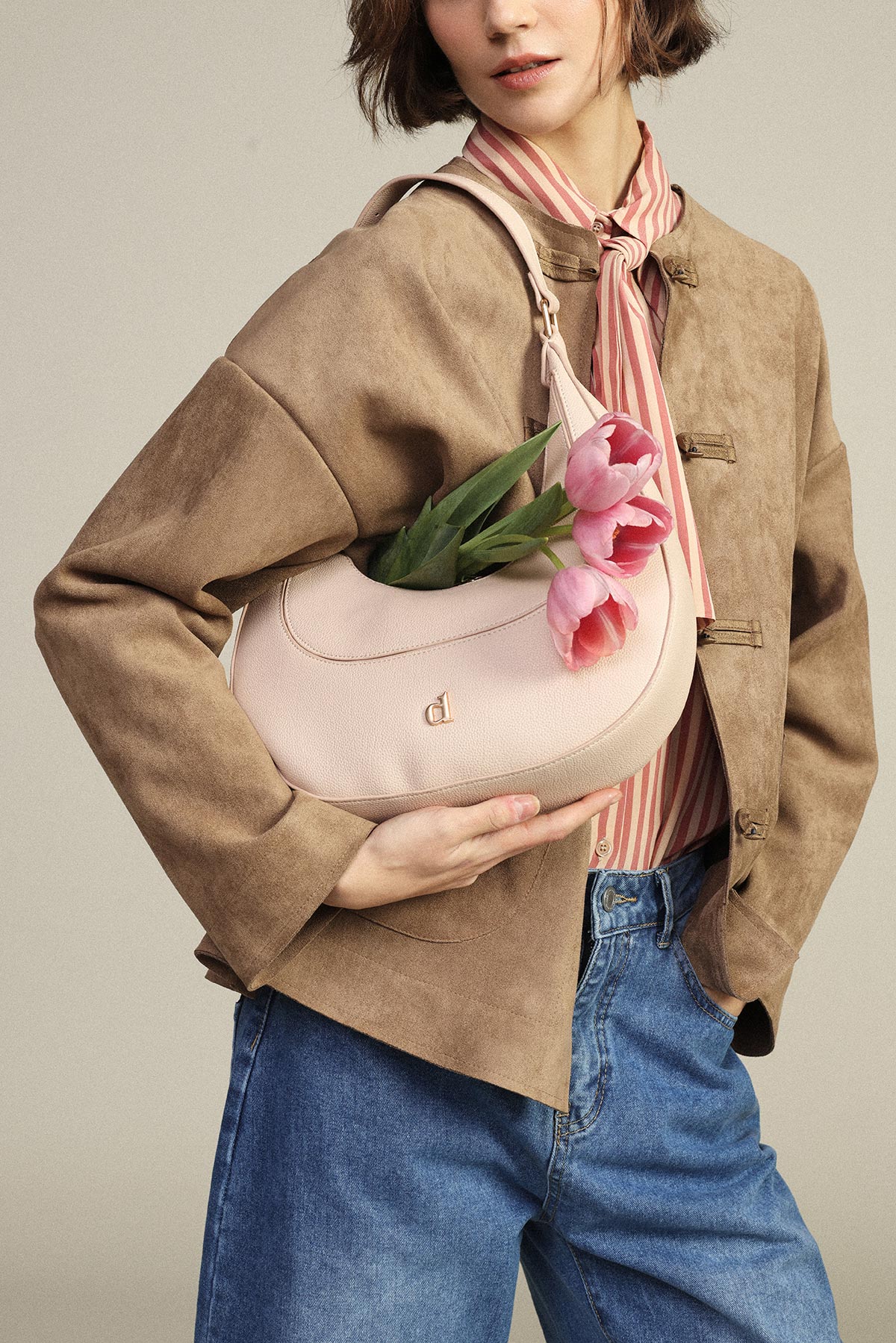 Woman in brown suede jacket and striped blouse holding a pale pink shoulder handbag with pink tulips, wearing blue jeans