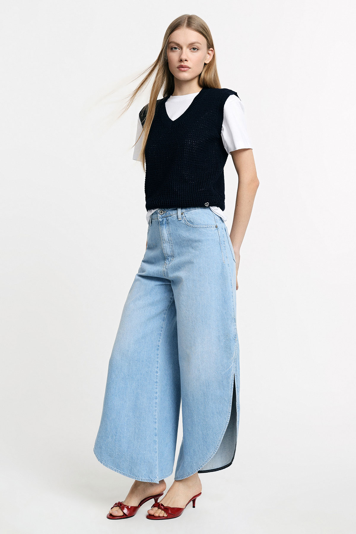 Model wearing black knitted vest over white tee, light‑blue wide‑leg cropped jeans with side slits and red heeled sandals