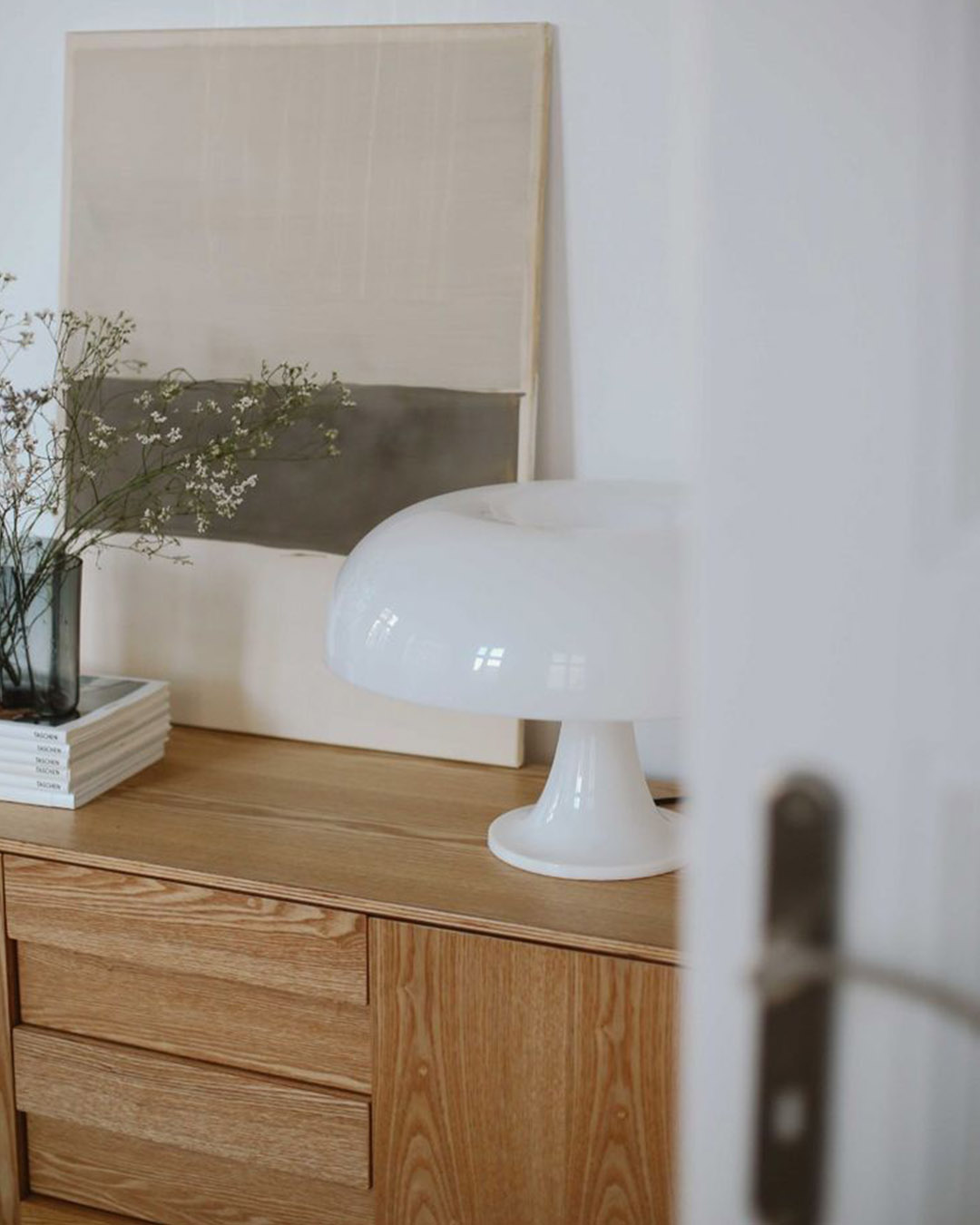 Minimal sideboard with oak drawers, white mushroom table lamp, abstract canvas, glass vase of gypsophila (baby's‑breath) and stacked books